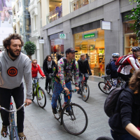 A group of cyclists from the 2010 Kritiska masa rides through a modern indoor shopping space. The cyclists wear varied attire: one in a gray hoodie with an orange emblem, and another in a colorful, patterned outfit. Shoppers and storefronts, including "The Body Shop," are visible in the background.