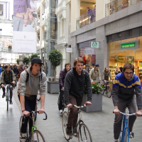 Three young men are riding bicycles through an indoor shopping mall. The mall is bustling with pedestrians, and stores like The Body Shop are visible. In the background, a large vertical banner displays the words "Galleria Center," featuring an image of two women with stylish attire, reminiscent of a Kritiska masa event from 2010.