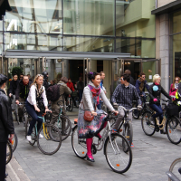 A group of people, dressed in casual and warm clothing, are riding bicycles through a glass-covered pedestrian area during Kritiska Masa 2010. Some cyclists wear helmets, and one person is dressed in a reflective yellow vest. The background shows a modern building with glass windows and doors, and various pedestrians are visible.
