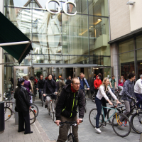 A group of cyclists are riding their bikes on a cobblestone path in front of a modern glass building with the letters "GC" displayed above the entrance. The lively scene resembles the Kritiska masa event from 2010, as pedestrians in various attires watch and add to the bustling atmosphere.