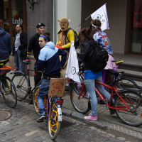 A group of people are gathered on a cobblestone street with their bicycles. One person, wearing a bear costume and holding a flag, stands out. The flags on two of the bicycles display a bicycle symbol. They appear to be participating in the 2010 Kritiska Masa bike event or rally near a building entrance with stairs.