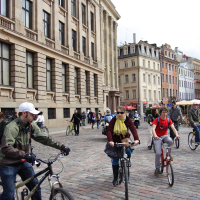 A group of people ride bicycles on a cobblestone street in an urban setting with historic buildings, reminiscent of a 2010 Kritiska masa event. Some cyclists wear helmets and casual clothing, while others don scarves and sunglasses. The sky is partly cloudy, and more pedestrians and cyclists are near a red vendor cart in the background.