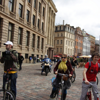 A group of cyclists, part of the 2010 Kritiska Masa, ride on a cobblestone street lined with historic buildings. One cyclist in a yellow scarf and brown jacket rides in the center, while another in a red shirt is on the right. A police officer on a motorcycle and a few pedestrians are in the background. The sky is overcast.