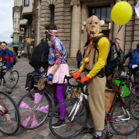 Two cyclists, dressed in colorful and quirky outfits, stand with their bikes in a cobblestone plaza in front of an old building. One cyclist wears a tiger mask, yellow jacket and holds a yellow balloon. The other is clad in a mismatched ensemble with magenta pants. More cyclists from the 2010 Kritiska Masa gather in the background.