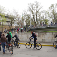 A group of people on bicycles are riding and walking in an urban setting near a gray bridge. The background features leafless trees and a brick building. Some cyclists head up a ramp while others remain at the bottom, evoking memories of the 2010 Kritiska masa cycling event or gathering.