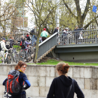 A group of people with bicycles are gathered on and around a pedestrian bridge in an urban park area. Some individuals are walking their bikes up a staircase, while others stand chatting. Trees with early spring foliage surround the scene, embodying the spirit of Kritiska masa. Two women in the foreground converse.