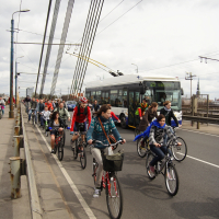 Cyclists ride on a busy bridge next to a moving bus and cars, reminiscent of Kritiska masa 2010. The group includes men, women, and children, some wearing helmets and casual clothing. Overhead wires and streetlights line the bridge, and the sky is overcast. The scene conveys active transportation amidst urban traffic.