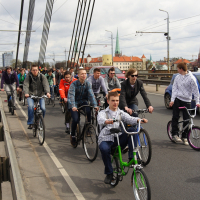 A large group of people, mostly young adults, are riding bicycles along a designated bike lane on a bridge. The cyclists appear to be participating in an organized event akin to the Kritiska masa of 2010. Overhead wires and city buildings with red rooftops and steeples are visible in the background under a cloudy sky.