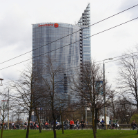 A modern glass skyscraper with the red "Swedbank" logo on top is in the background. In the foreground, a large group of cyclists, part of the Kritiska masa movement, rides along a road lined with leafless trees. Overhead power lines stretch across the cloudy sky in this 2010 urban scene.