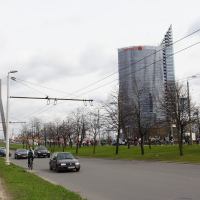 A cityscape featuring a tall glass skyscraper in the background with a distinctive angular design at the top. In the foreground, there is a busy street with cars, cyclists from Kritiska masa, and pedestrians. A modern bridge with cables spans the road, and leafless trees line the street under a cloudy sky, evoking 2010 vibes.