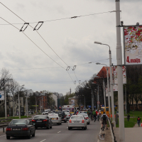 A busy urban street with several cars and a bicyclist, reminiscent of Kritiska Masa 2010, dominates the foreground. Tall lampposts and banners, one reading "4.maijs," line the street. Buildings and trees are visible in the background under a cloudy sky, while overhead power lines crisscross above.