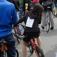 A group of people, some on bicycles, gathered in daylight. A girl in a striped shirt rides a small red bicycle with "Masa Kritiska 2010" pinned to her back. The crowd includes various people standing and interacting around her, with many holding or riding bicycles.