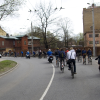 A large group of cyclists ride together on a curved urban street, surrounded by buildings and leafless trees, reminiscent of the 2010 Kritiska masa events. The cyclists range in age and attire, indicating casual participation. A motorcyclist follows behind them. The day appears overcast, and the street has minimal vehicular traffic.