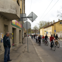 A man stands on the sidewalk near an old building with graffiti and a sign reading "Kinga-Linija" in a foreign language. Multiple cyclists, like those from the Kritiska masa 2010 event, ride down a narrow street under cloudy skies. The street is lined with more old buildings and street signs, and there is a small crowd visible in the distance.