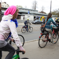 People of various ages are riding bicycles down a city street, reminiscent of a Kritiska Masa event. The closest cyclist, wearing a pink beanie and white hoodie, is on a green bike. Other cyclists are ahead, including one in a teal sweater with a backpack. Buildings, parked cars, and trees line the background.