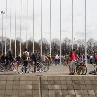 A bustling outdoor scene captures numerous people standing around bicycles, reminiscent of Kritiska Masa 2010. The individuals are in casual clothing, some prepping to ride. The area is paved, and tall flagpoles without flags stand in the background. Bare trees and a cloudy sky frame the setting beautifully.