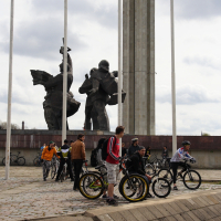 A group of people with bicycles gathers in front of a large, tall monument featuring two abstract statues against a cloudy sky. The scene, reminiscent of Kritiska masa 2010, appears to be in a public plaza with concrete paving and flagpoles. The cyclists are engaged in conversation or preparing their bikes.