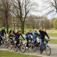A group of cyclists, including four adults and two children, ride along a paved path in a park during a 2010 Kritiska masa event. The lead male cyclist waves to the camera. Bare trees line the path, and a building with large windows is visible in the background. The sky is overcast, with grass and budding trees surrounding the path.