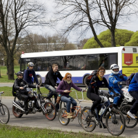 A group of people wearing helmets and casual clothing ride various bicycles and a tandem bike on a paved path beside a grassy area with trees. Behind them, a white bus with purple and yellow accents is in motion. The overcast scene, reminiscent of the 2010 Kritiska masa event, conveys an outdoor activity.