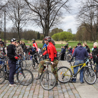 A large group of cyclists gather in a park, chatting and preparing for a ride. They are dressed in casual and sporty clothes, some wearing helmets. The area, paved with cobblestones and surrounded by leafless trees, evokes early spring. The mood is lively and social, resembling a 2010 Kritiska masa event.