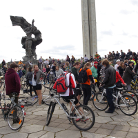 In 2010, a large group of people with bicycles gathered in an open plaza featuring a tall sculpture in the background, reminiscent of a Kritiska masa event. Many individuals are standing and conversing, with some sitting on their bikes. The atmosphere appears lively and social, with a mix of cloudy skies overhead.