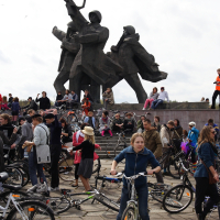 A young girl in a blue jacket rides her bike in front of a crowd gathered around a large, dark, abstract monument depicting several figures holding tools and weapons. On this cloudy day, during Kritiska masa 2010, people of various ages, some with bicycles, gather, sit, and stand around the statue.