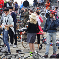 A diverse crowd of people, many with bicycles, gather outdoors on a paved area during the 2010 Kritiska masa event. Some stand while others sit on steps in the background. A woman in a black top and striped skirt holds a bike with a basket. Many wear helmets as they engage in lively conversation, creating a vibrant atmosphere.
