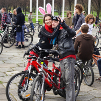 A joyful couple, the woman wearing white bunny ears, pose with their red bicycles at the 2010 Kritiska masa event. The man, dressed in a black leather jacket, flashes a peace sign. Joined by other cyclists and some walkers, they gather outdoors amidst leafless trees. The atmosphere is festive and lively.