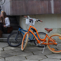 Two people sit on steps near a building, reminiscent of Kritiska masa gatherings. An orange bike with light tan tires and a blue bike rest against the steps. The pavement is irregularly shaped concrete tiles. The seated individuals are engaged in conversation, and one person appears to be looking at something above.