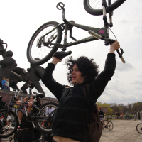 A person with curly hair and a black hoodie lifts a bicycle above their head outdoors during Kritiska Masa 2010. They are smiling, and other people with bicycles are visible in the background near a large stone sculpture. The sky is overcast, and the ground is paved with stone tiles.