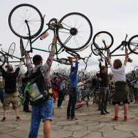 A diverse group of cyclists, part of the 2010 Kritiska masa event, energetically hold their bicycles above their heads in a celebratory manner. They stand on a paved area with a cloudy sky above, demonstrating enthusiasm and community spirit. Trees can be seen in the background.