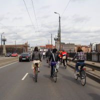 A group of cyclists rides across a wide bridge alongside a single lane of car traffic, under a cloudy sky. The cyclists, part of a Kritiska masa event, are dressed in casual attire. The bridge features tram tracks and lamp posts, with historic buildings and a church steeple visible in the skyline.