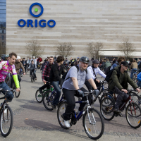 A large group of people participate in a Kritiska masa event, riding bicycles outdoors near a commercial building with the "ORIGO" logo prominently displayed. The cyclists, wearing casual clothing and bike helmets, move in different directions. The background includes other pedestrians, a building wall, and trees.