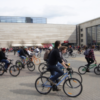 A bustling scene of numerous people riding bicycles in an urban square depicts the Kritiska masa event of 2010. The background shows a modern, large building labeled "Origo." The crowd is diverse, including adults and children with varied bike styles. The sky is overcast, and the atmosphere appears lively and active.