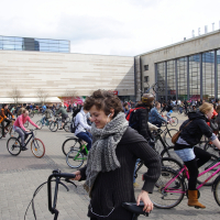 A bustling urban square filled with numerous cyclists riding in different directions. In the foreground, a woman in a gray scarf and dark jacket is walking her bicycle with a smile on her face. Behind her, many people are also on bicycles, reminiscent of the Kritiska masa event from 2010, with buildings visible in the background.