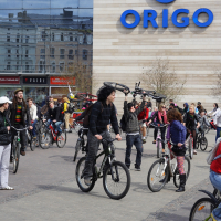 A large group of people on bicycles gathered in front of a modern building with a sign that reads "ORIGO." Reminiscent of the 2010 Kritiska masa events, the cyclists are of various ages, wearing casual clothing. The area is paved with riders and those standing with their bikes. Reflective windows show additional buildings.