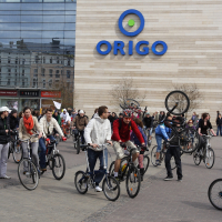 A group of cyclists is gathered in front of the Origo shopping center, reminiscent of the Kritiska Masa event in 2010. Some individuals are performing stunts, including a person mid-wheelie. Numerous spectators are watching the event. The building features a large glass facade and beige walls with the blue "ORIGO" logo prominently displayed.