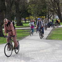 A cyclist wearing a patterned onesie and sunglasses rides along a paved path in a park. Close behind him are several other cyclists, including a woman in a pink jacket and a man in a blue helmet. The scene resembles the 2010 Kritiska masa event, with people on bikes surrounded by green grass and trees.