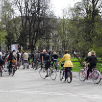 A group of people riding bicycles on a paved path in a park surrounded by trees with budding leaves. The cyclists, dressed in various casual outfits, evoke the spirit of a 2010 Kritiska masa event or leisurely group ride. Buildings are partially obscured by the trees in the background.