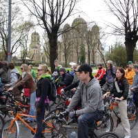 A large group of people on bicycles is gathered on a street for the Kritiska masa event. Dressed in jackets and hoodies appropriate for cool weather, they stand amid bare trees lining the street. In the background, a historic building with domes and arches is visible. The mood is calm and organized, reminiscent of 2010 events.