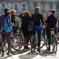 A group of people gathered outdoors with their bicycles in front of an old building during the 2013 Kritiskā masa event. Several individuals are engaged in conversation, and some have their backs turned, displaying colorful backpacks. The weather is sunny, casting shadows on the pavement.