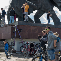 A group of people gathers around large, abstract sculptures. Several are taking photos, and some are riding or standing with bicycles. A tall, unusually designed bicycle stands out. The scene is casual, reminiscent of Kritiskā masa 2013, with individuals of various ages, including children, engaged in leisure activities.