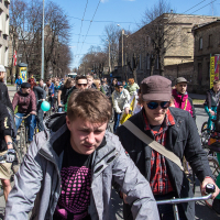 A bustling crowd of cyclists pedals down a city street lined with bare trees and brick buildings. The riders, clad in various outfits including jackets and hats, navigate the narrow road together. Colorful signs and street art add vibrancy to the scene under a clear blue sky during Kritiskā masa.
