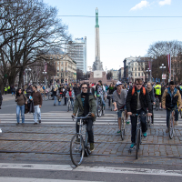 A large group of cyclists rides on a cobblestone street near a monument with a statue on top. Leafless trees and buildings surround the area under a clear sky. Some cyclists wear sunglasses and backpacks, while pedestrians are seen in the background. This 2013 event is part of Kritiskā masa.