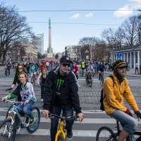 A large group of cyclists, including men and women of different ages, ride during the 2013 Kritiskā masa rally on a city street. In the background is a tall monument and buildings with modern and classic architecture. Trees without leaves line the street, and the sky is partly cloudy with blue patches.