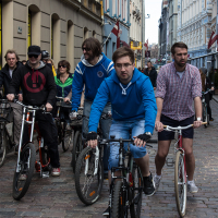A group of people is riding bicycles on a cobblestone street in an urban area, reminiscent of the 2013 Kritiskā masa. The riders, wearing casual clothing including hoodies, jackets, and shorts, are focused ahead as they navigate the narrow, building-lined street. The background shows a mixture of colorful buildings and pedestrians.