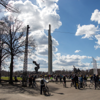 A large group of cyclists gather under a clear, sunny sky for the 2013 Kritiskā masa. Bare trees line the sides of the path, and statues flank a tall, narrow monument in the center distance. Puffy clouds float in the blue sky, creating a backdrop for the lively scene of people and bicycles.