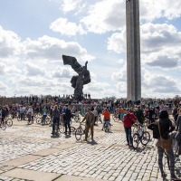 A large group of people, many with bicycles, gather at a public square on a partly cloudy day during the 2013 Kritiskā masa. Dominating the background is a tall, vertical monument and a dynamic sculpture of a figure holding a flag. The area is paved with light-colored stones and bricks.