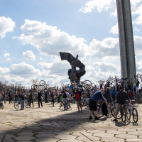 A large group of cyclists gather in a plaza on a sunny day, lifting their bicycles in the air in front of a towering sculpture. The sky is dotted with clouds, and many of the participants are dressed in casual clothing. This 2013 event marks Kritiskā masa, held on the stone-tiled plaza with a raised platform in the background.