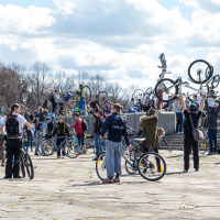 A large group of people gathered outdoors, each lifting their bicycles above their heads in celebration. The sky is partly cloudy, and leafless trees are visible in the background. The crowd spans a wide open area with paved ground, capturing the festive and communal spirit of Kritiskā masa 2013.
